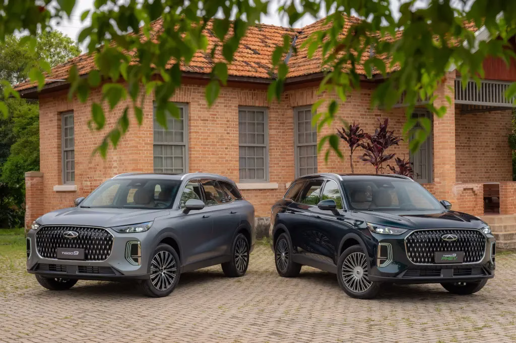 Dois CAOA Chery Tiggo 9, um cinza e outro verde-escuro, estacionados em frente a um prédio de tijolos