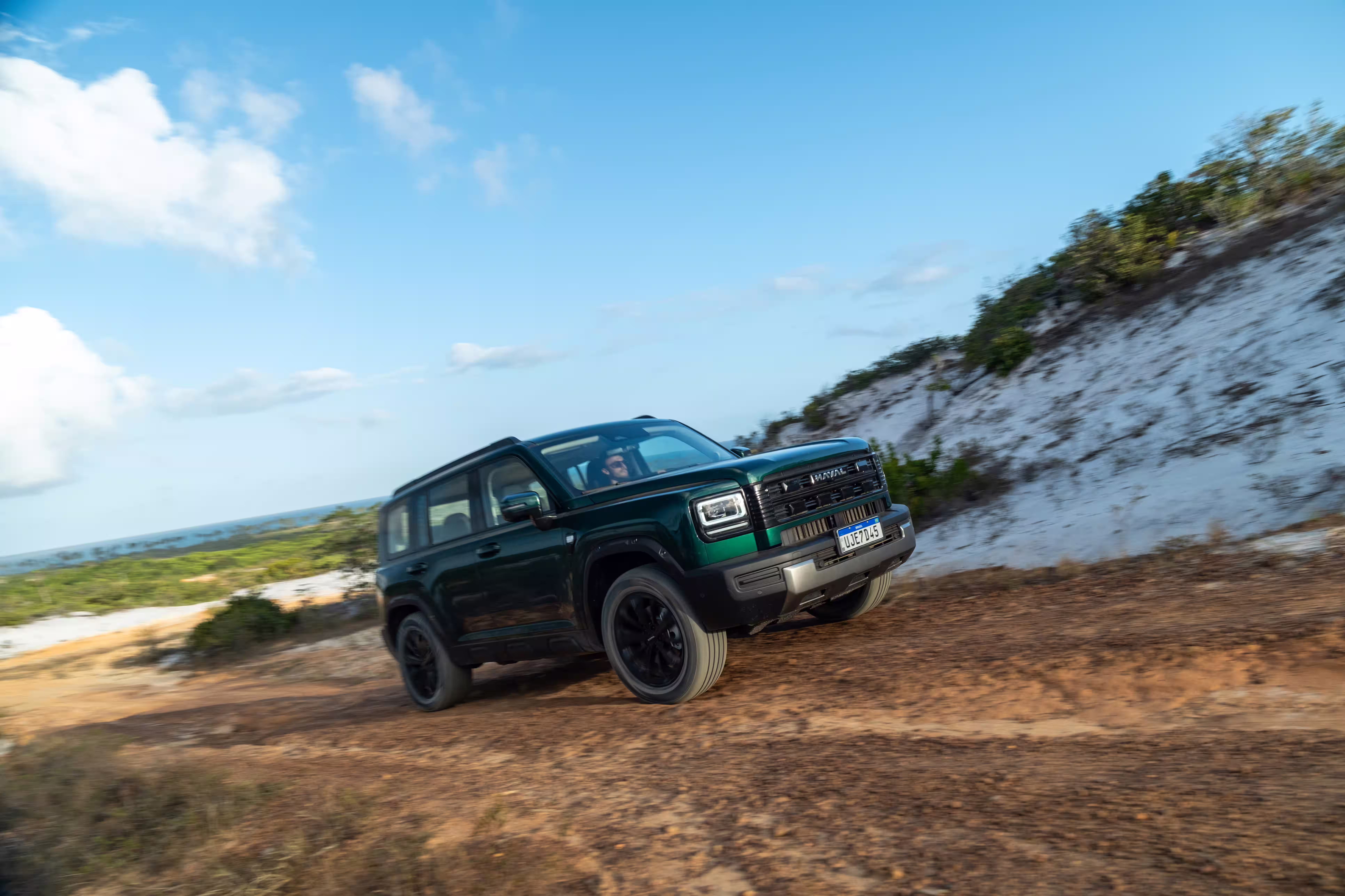 Foto de divulgação: Haval H7 verde de lado e de frente numa estrada de terra junto a vegetação e areia.