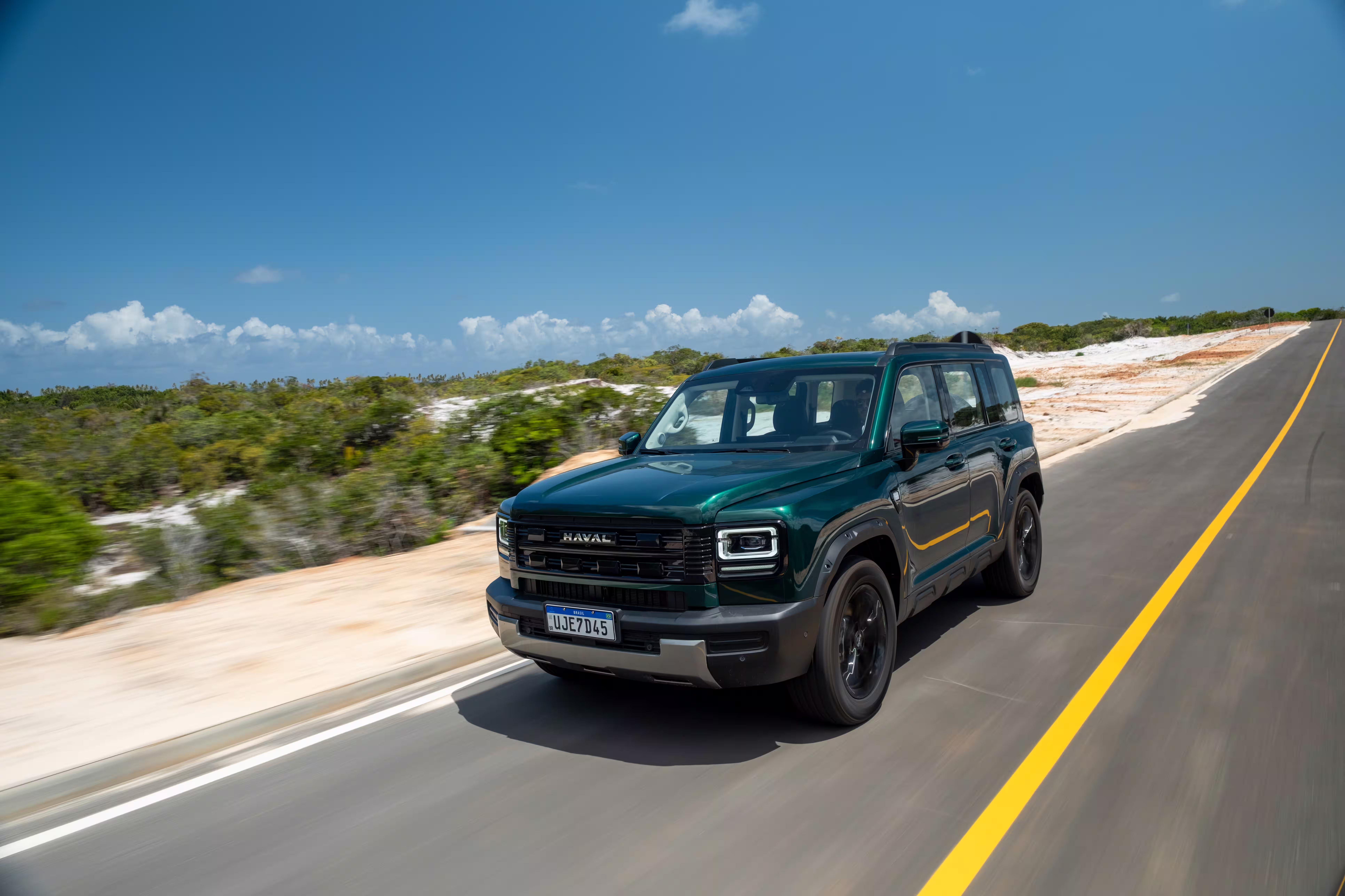Foto de divulgação: Haval H7 verde em três quartos dianteiros numa via asfaltada junto a areia e vegetação baixa.