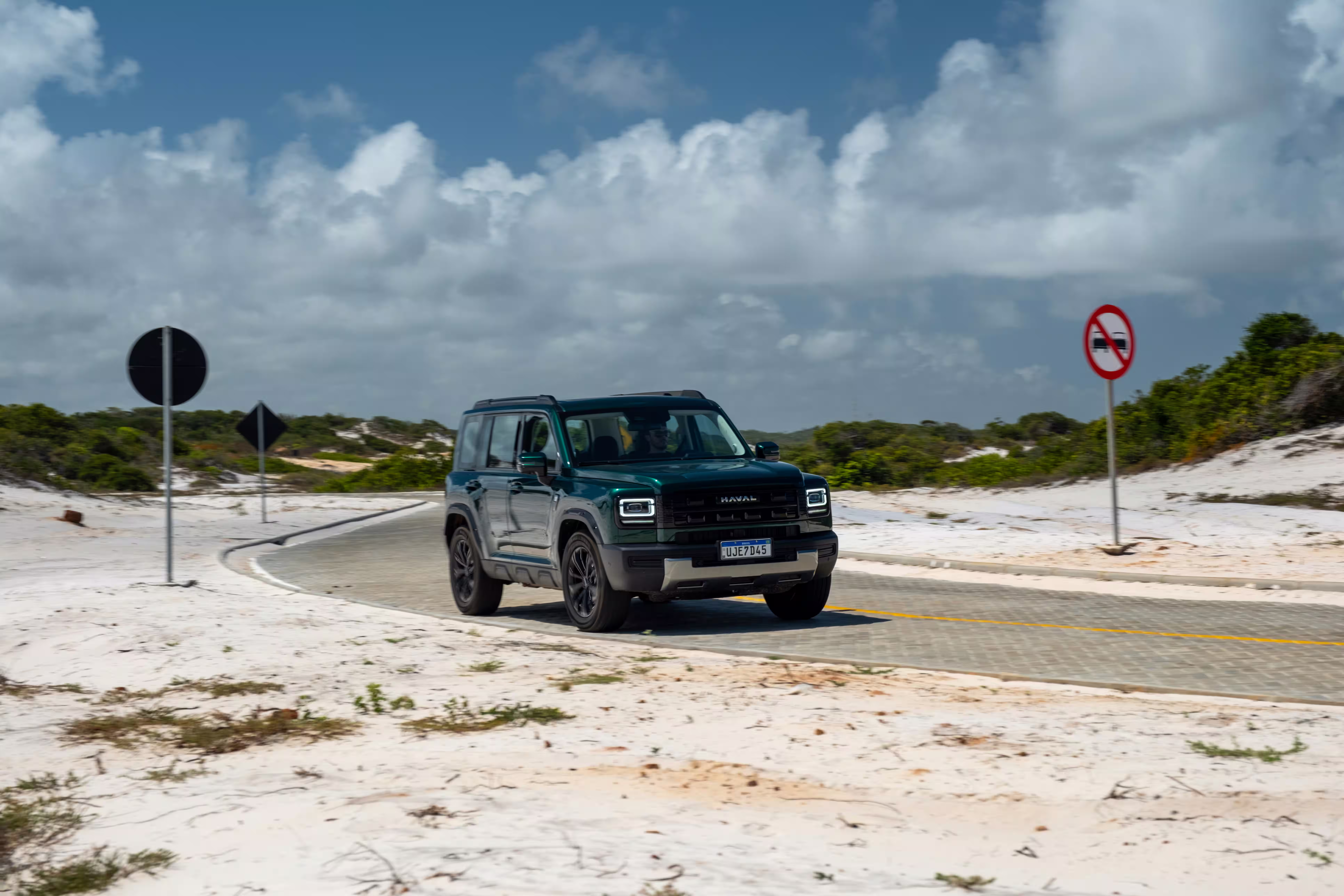 Foto de divulgação: Haval H7 verde contornando uma curva de asfalto entre dunas claras e placas viárias.