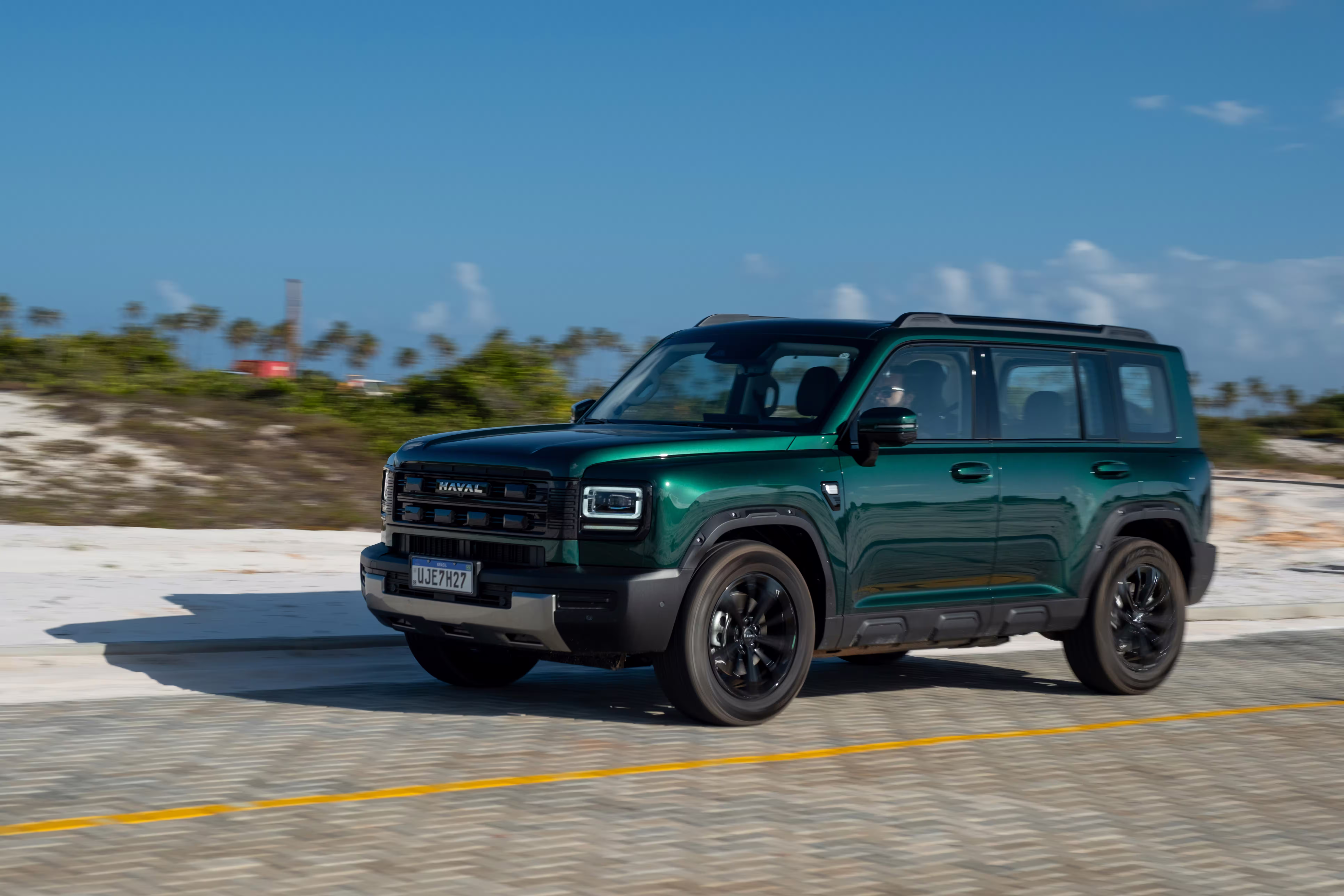 Foto de divulgação: Haval H7 verde em três quartos dianteiros num trecho de asfalto junto a areia clara.