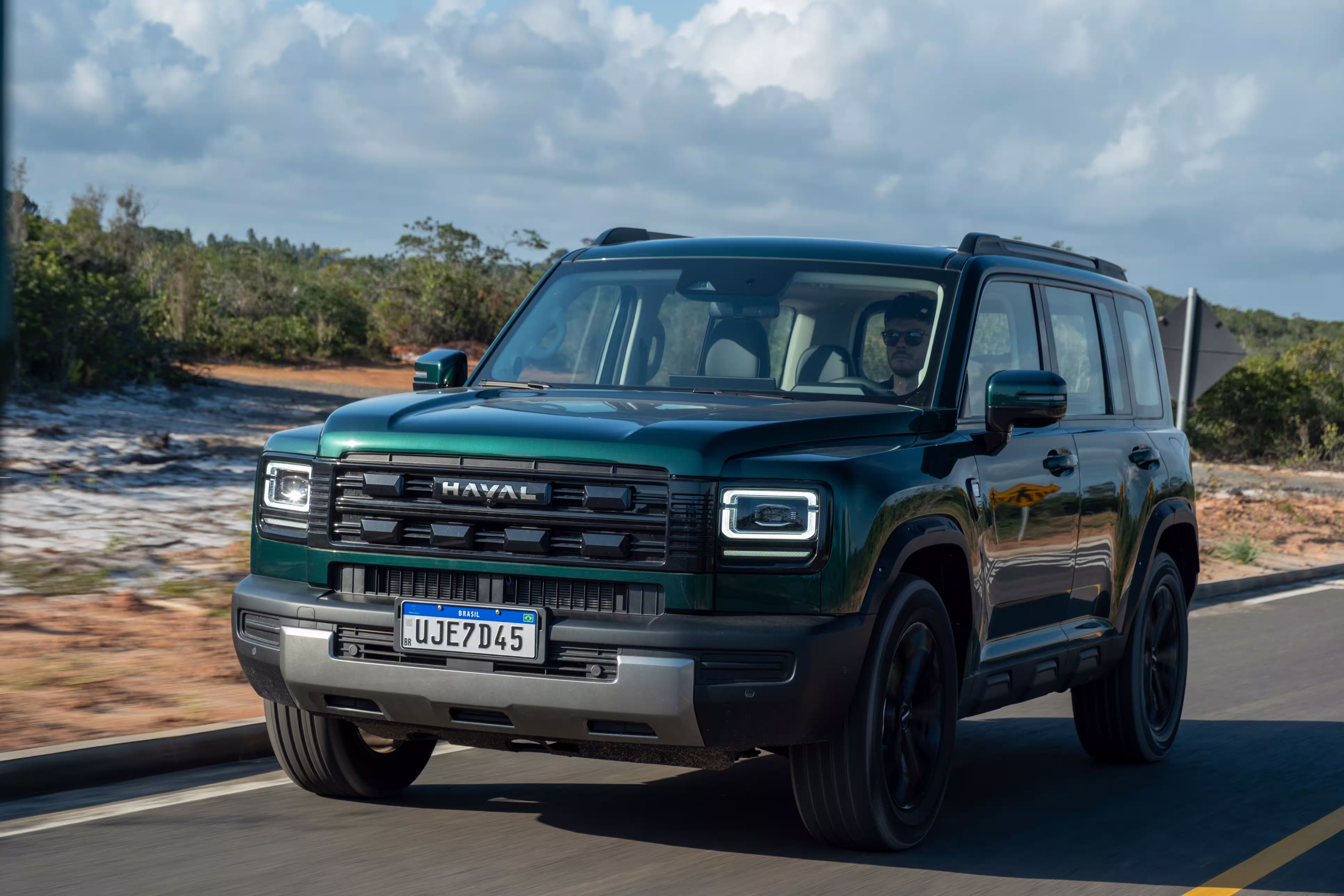 Foto de divulgação: Haval H7 verde em três quartos dianteiros sobre uma via de asfalto, sob céu com nuvens.