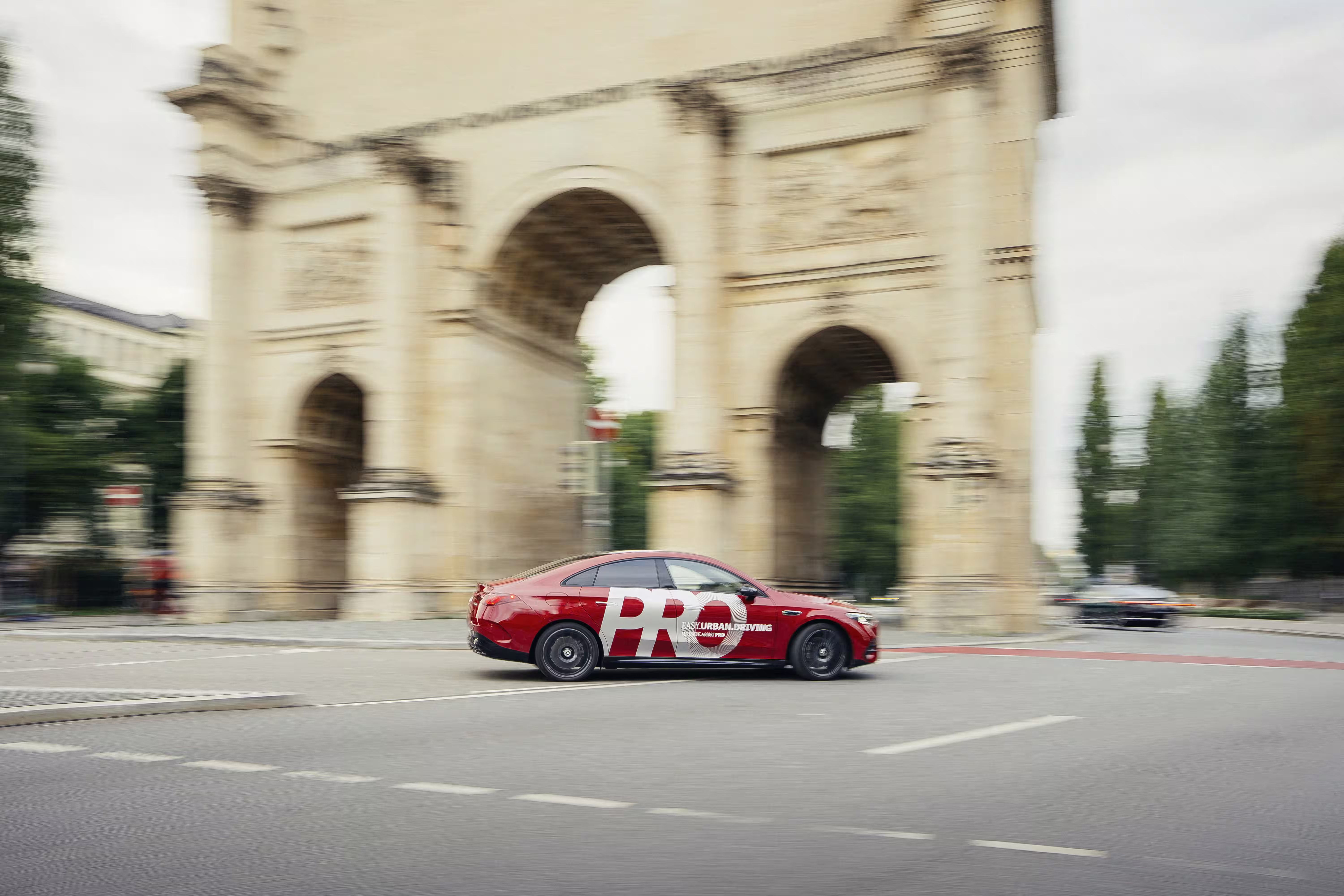 Mercedes-Benz vermelho com grafismo PRO passando diante de um arco monumental em uma rua