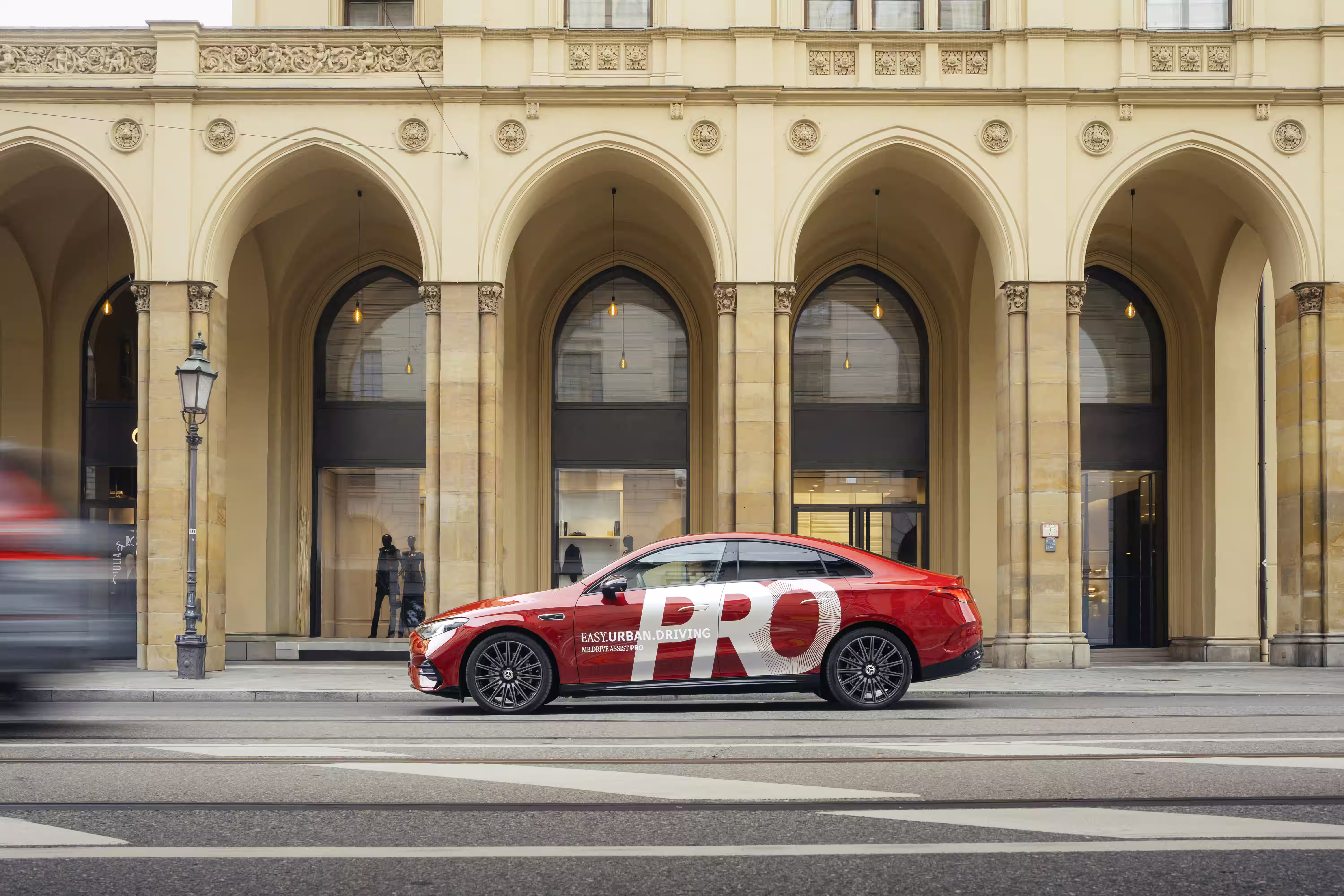Mercedes-Benz vermelho com identificação MB.DRIVE ASSIST PRO visto de perfil diante de uma fachada com arcos